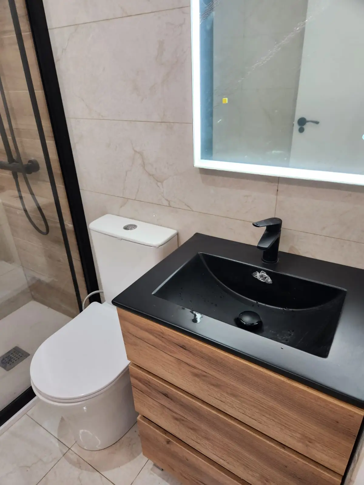 Reforma de baño con lavabo negro mate sobre mueble de madera y espejo retroiluminado en Barcelona