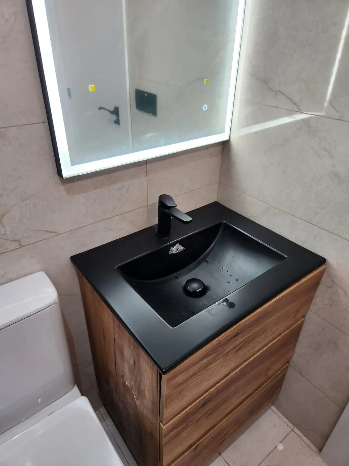 Reforma de baño con lavabo negro mate sobre mueble de madera y espejo retroiluminado en Barcelona