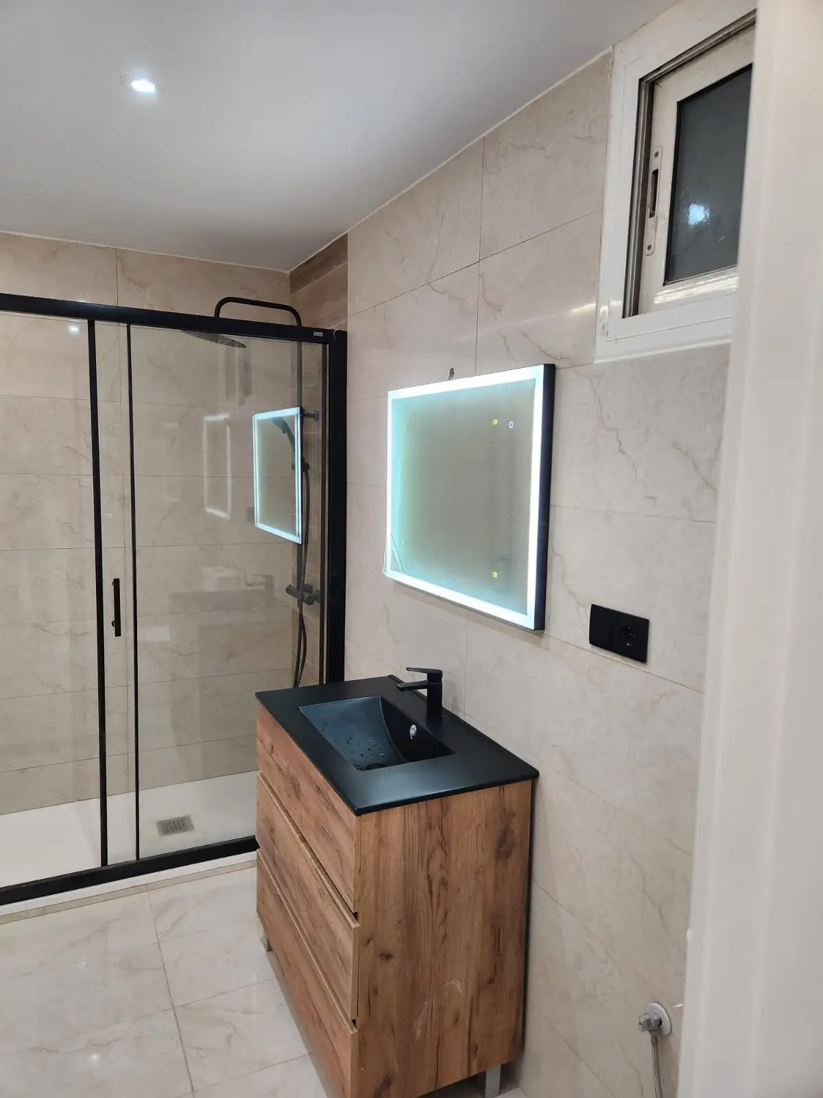 Reforma de baño con mueble de madera, lavabo negro y espejo retroiluminado en Barcelona